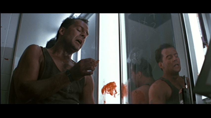 Medium close-up of McClane contemplating the piece of glass he has just removed from the left half of the frame, with his reflection in a mirror occupying the right third of it