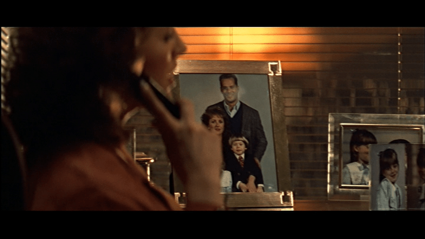 Holly, out of focus in the left side of the frame, talks on the phone in front of a framed family photo