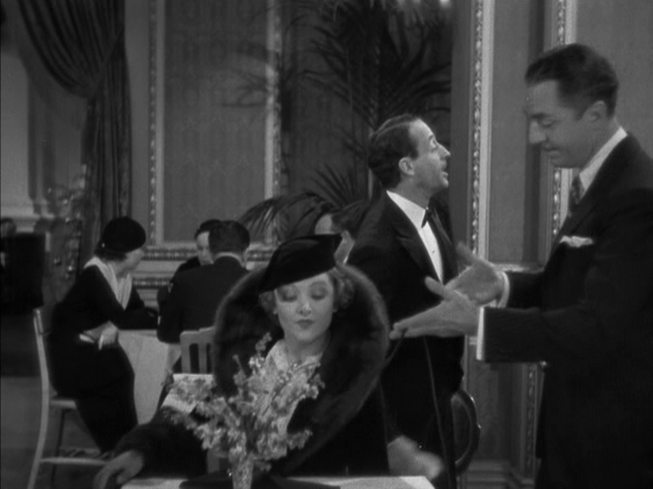 Medium shot of Nick gesturing at a table from the right side of the frame which Nora sits down at, centered in the bottom half of the mage, as a waiter calls for two cocktails in the background