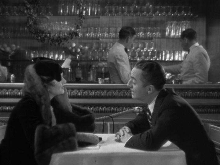 Medium shot of Nick on the bottom-right side of the frame sitting across a table from Nora on the bottom-left side of the frame as they talk against a backdrop of glassware and barmen