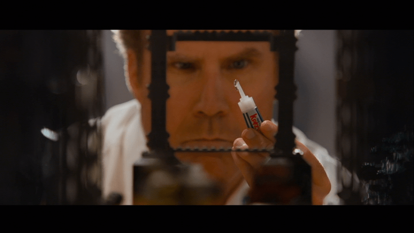 Close-up of Will Ferrell's The Main Upstairs holding a bottle of Krazy Glue which is in focus and he's slightly out of focus behind in a  frame within a frame created by one of his Lego creations