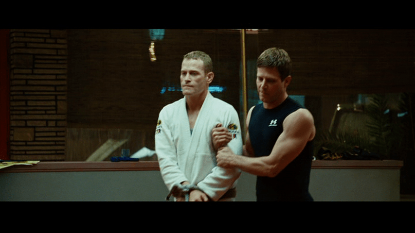 Medium close-up of Officer Joe being "taken to court" with his hounds bound by another fighter (Matt Cable)