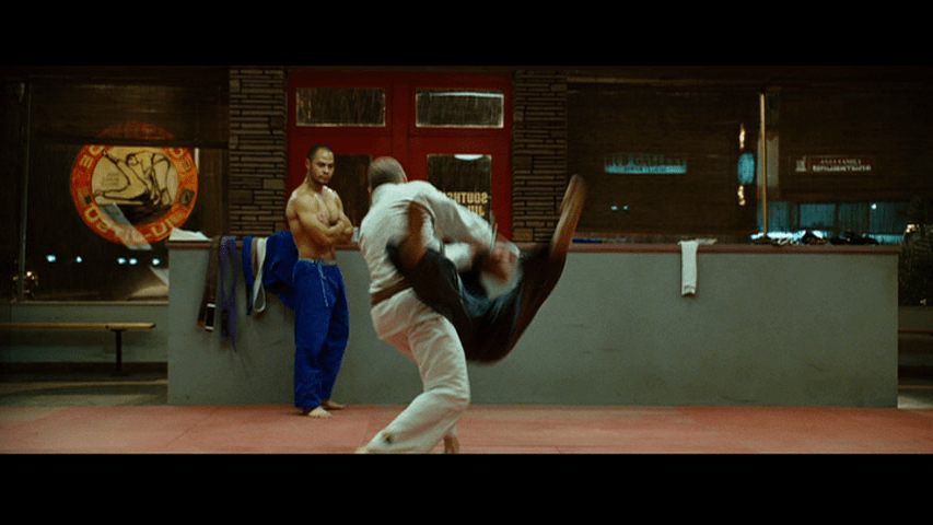 Long shot of Officer Joe executing a takedown as another instructor named Snowflake watches from the back of the dojo