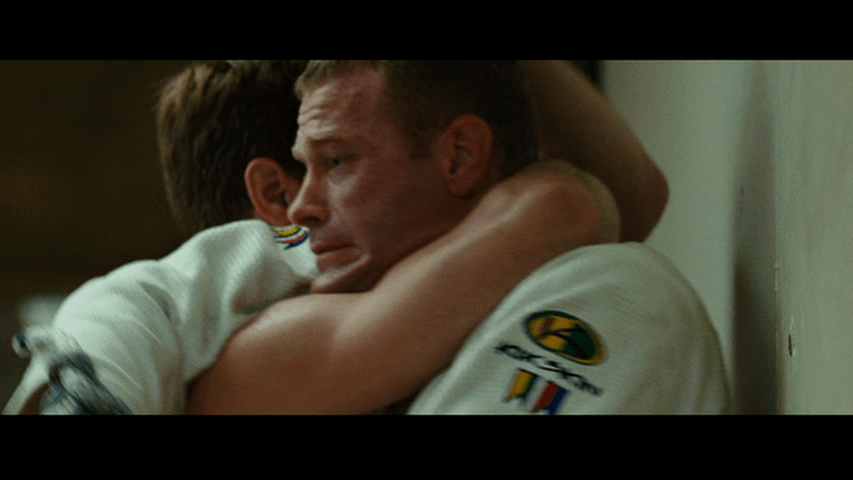 Close-up of Officer Joe centered in the frame, his back to the wall, with the other fighter's arm choking him into submission