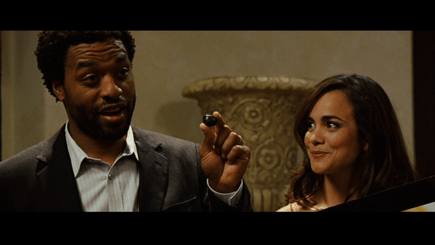 Close-up of a besuited Mike holding up a black rock that looks like one of the marbles used in his training method from the left half of the frame as his wife Sondra (Alice Braga) smiles at him from the right