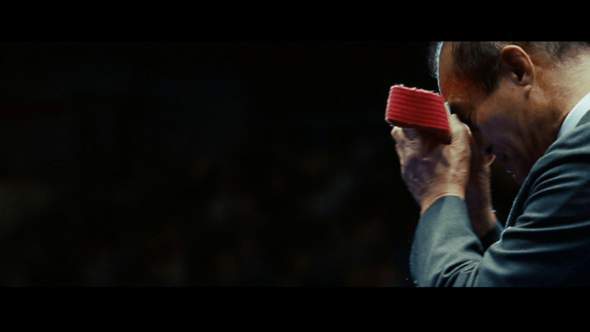 The Professor holds the red belt of the film's title up against his forehead against the same black background in the right third of the frame