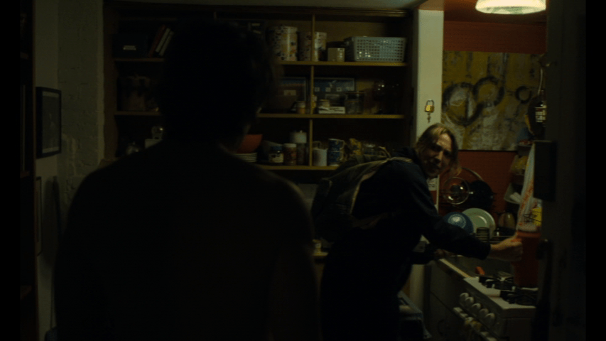 Medium shot of Alex, whose back is to the camera, talking to Jill, who is dressed in scrubs and preparing to leave for work, in a cramped kitchen