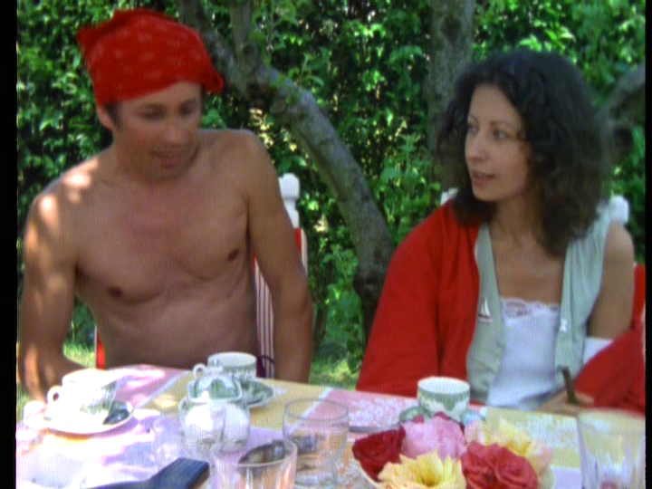 Medium shot of Delphine and a shirtless man sitting at an al fresco tea table on a sunny day