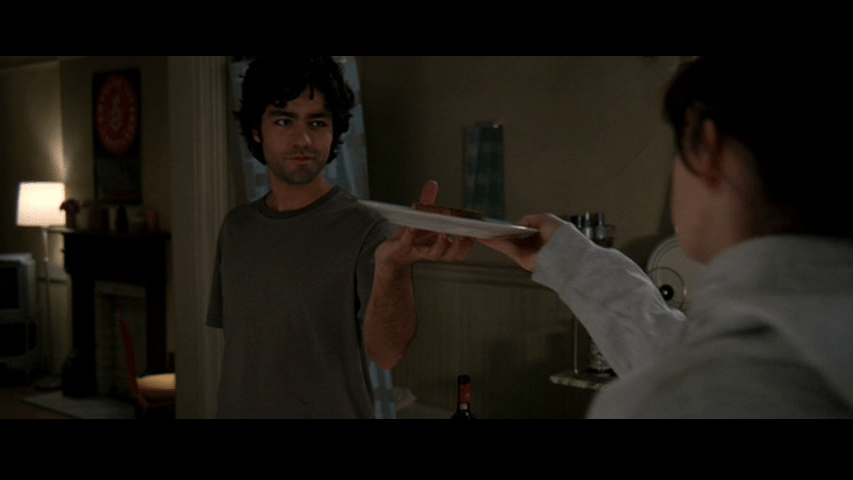 Medium shot of Nate handing Andy, who is out of focus in the right edge of the frame with her back to the camera, the sandwich he's been making on a plate