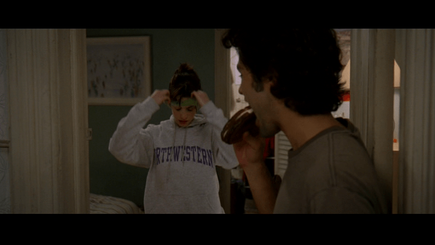 Medium shot of Nate eating the grilled cheese he made for Andy in the right foreground as she puts on a headband in the center of the frame