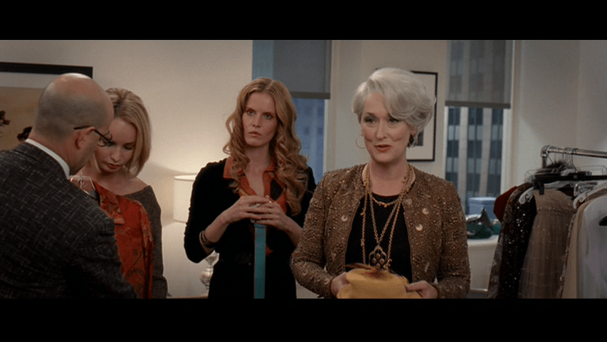 Medium long shot of Miranda chastising an offscreen Andy from the center-right of the frame while Nigel holds a jacket up in front of a model next to another Runway employee holding a belt