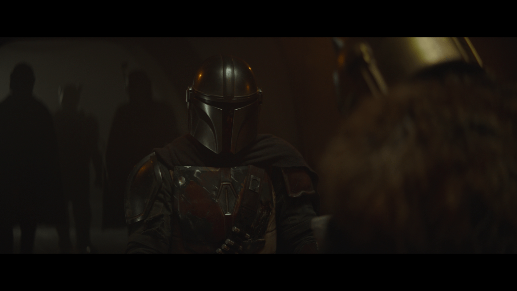 Medium close-up of Din Djarin sitting before the Armorer, who is out of focus in the foreground with her back to the camera, as a crowd of Mandalorians forms behind him