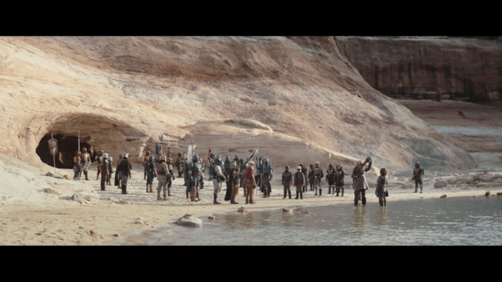 Long shot of the Armorer holding a helmet above an initiate as they both stand in water and a crowd of Mandalorians looks on from just outside the entrance to a cave