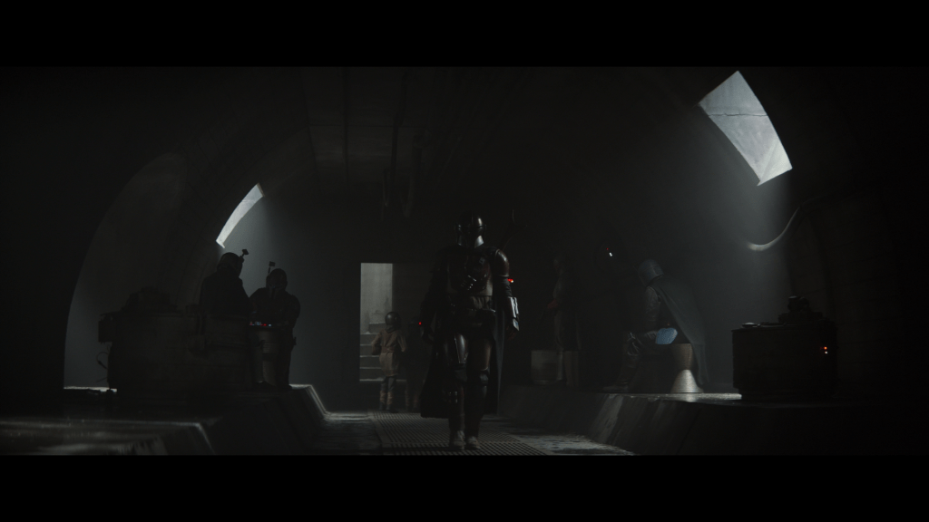 Long shot of the Mandalorian walking down a hallway flanked by other Mandalorians