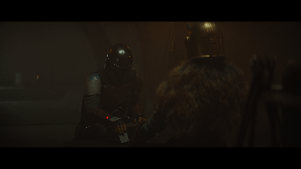 Medium long shot of Din Djaron placing an ingot of Beskar in front of the Armorer, who sits facing him with her back to the camera in the right foreground