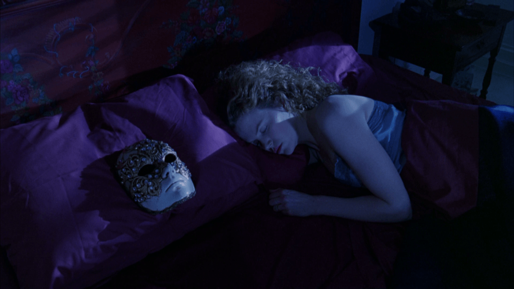 Medium POV shot of Nicole Kidman's Alice Harford from her husband's perspective, asleep in purple sheets with the mask he wore during his misadventures the previous night carefully positioned on his empty pillow