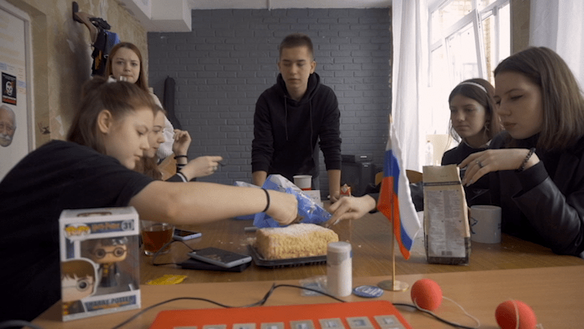 Medium shot of students gathered around a table in Pavel Talankin's office with a Harry Potter toy in the foreground