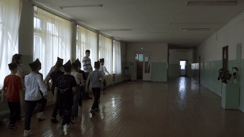 Medium long POV shot of Pavel observing students conducting marching drills in the hallway of his school