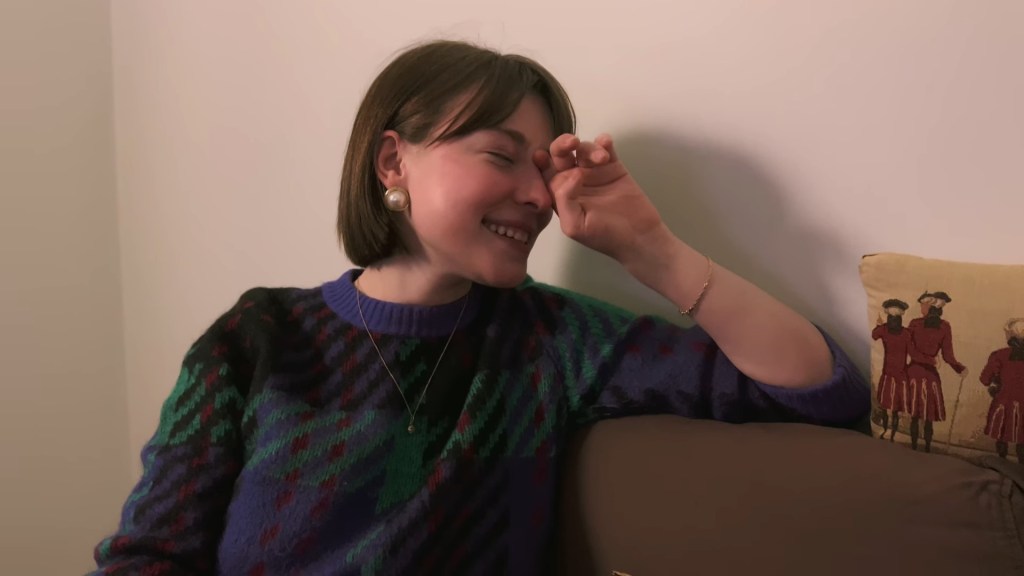 Medium close-up of Ksenia sitting on a couch laughing, but with a hand over one of her eyes and a fatalistic expression