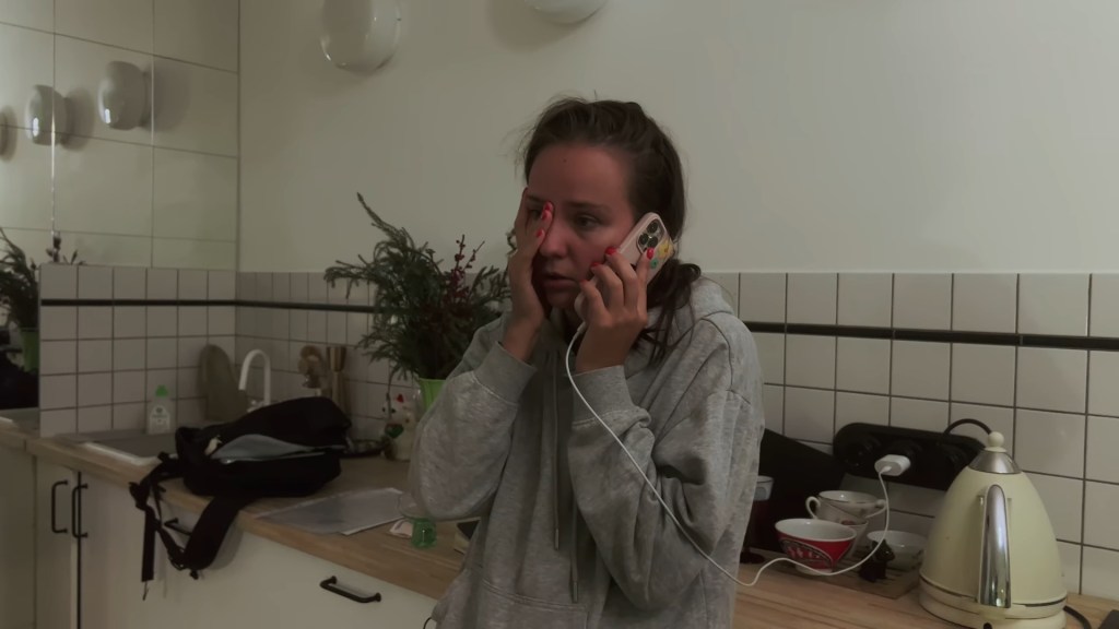 Medium shot of Olya listening to a voicemail in her kitchen with a worried expression