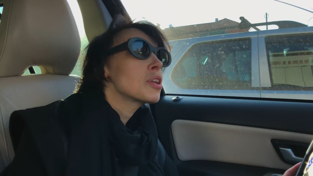 Medium shot of Anya craning her neck to look at something out the windshield of the car she's driving