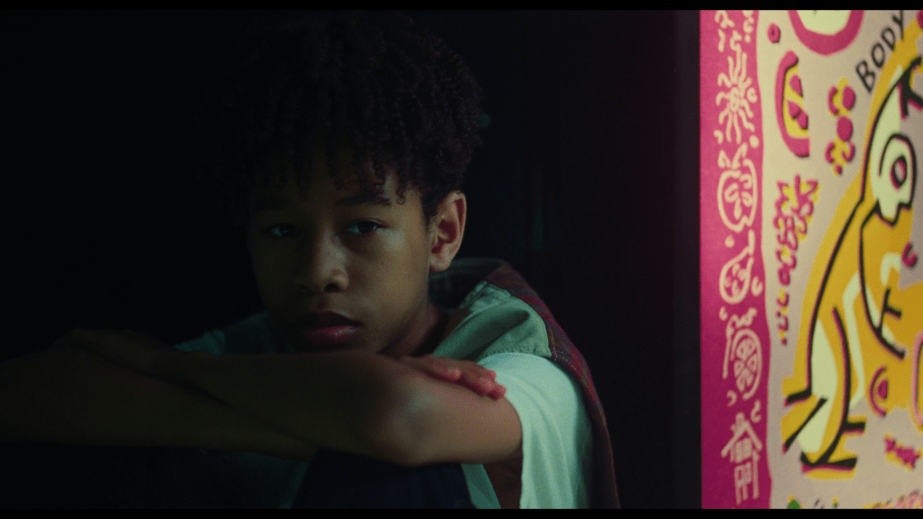 Close-up of Owen, arms crossed, sitting next to a Fruitopia machine which occupies most of the right third of the frame