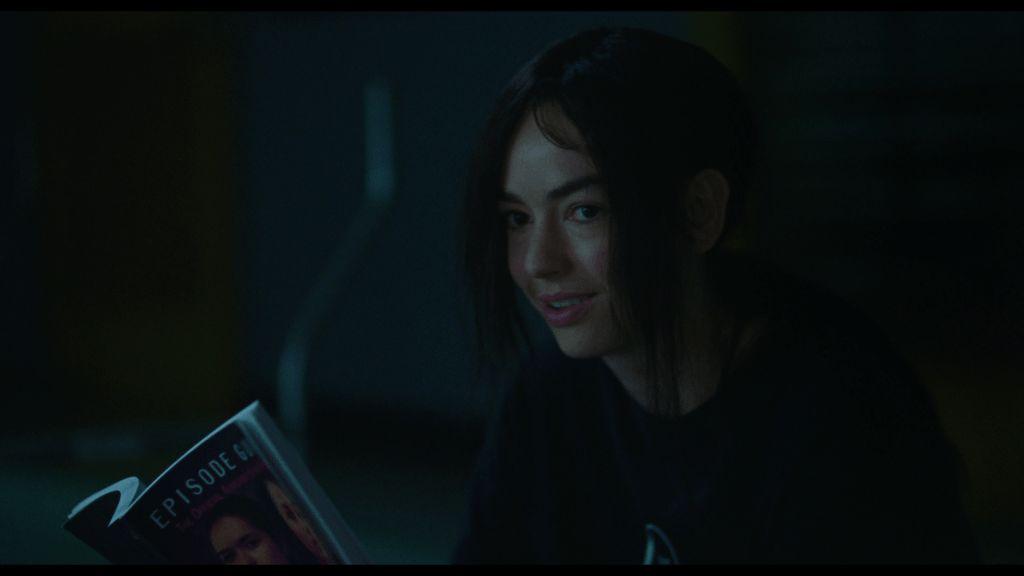 Close-up of Maddy smiling in disbelief, the Pink Opaque episode guide she's holding visible in the bottom-left corner of the frame