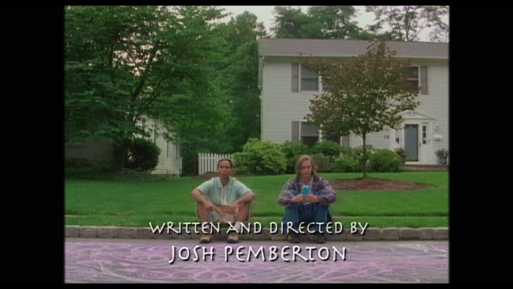 Long shot of two teenagers sitting on a curb eating popsicles with the credit "Written and Directed by Josh Pemberton" superimposed on top of them