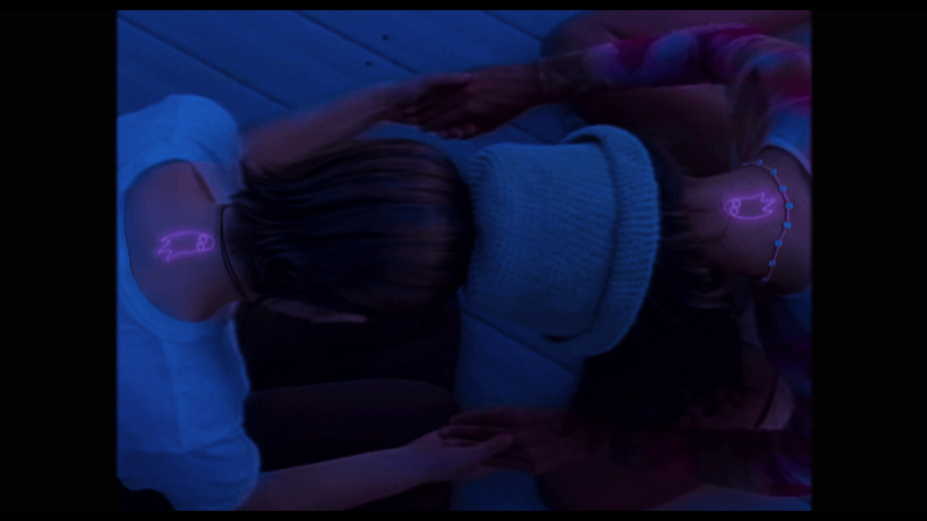 Overhead shot of Tara and Isabel clasping hands and putting their heads together while the ghost-shaped birthmarks they each have on the back of their neck glow pink