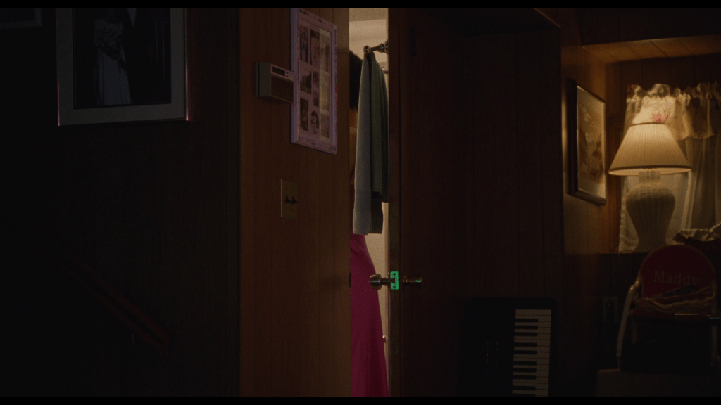 Medium long shot of Owen trying on a pink dress in Maddy's bathroom, just barely visible through a crack in the door