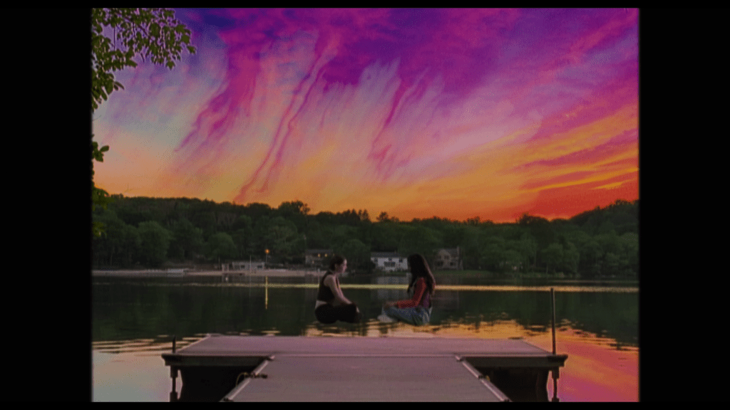 Long shot of Tara and Isabel floating above the dock at their sleepaway camp facing each other under a sky of outrageous pinks, purples, and oranges