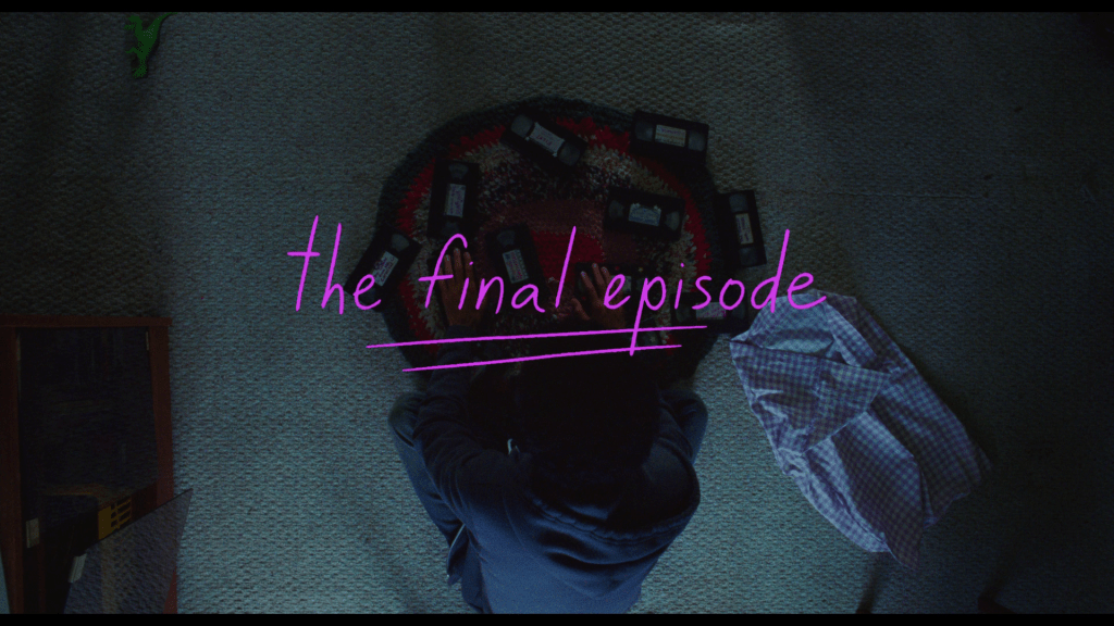 Overhead shot of Owen sitting in front of the pile of VHS tapes he has just dumped out of the gingham bag next to him under pink titles that read "The Final Episode"