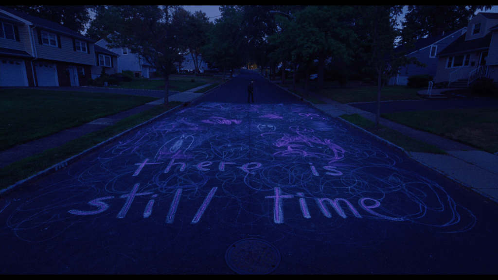Extreme long shot of Maddy looking back at the camera from the middle of a street upon which she has written in chalk, "there is still time."