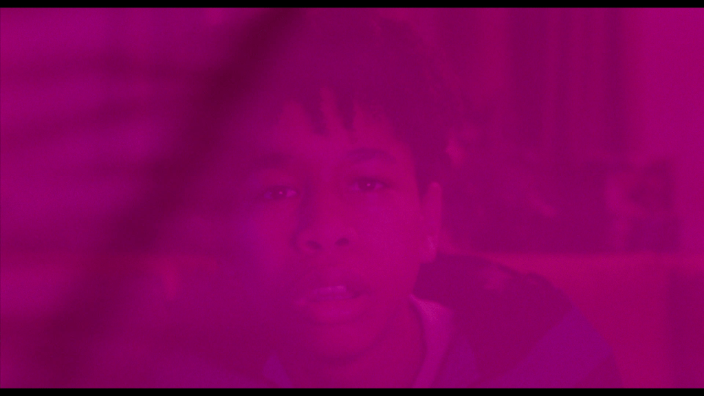 Close-up of Owen staring out at the camera as he watches TV in a shot drenched in pink because . . .