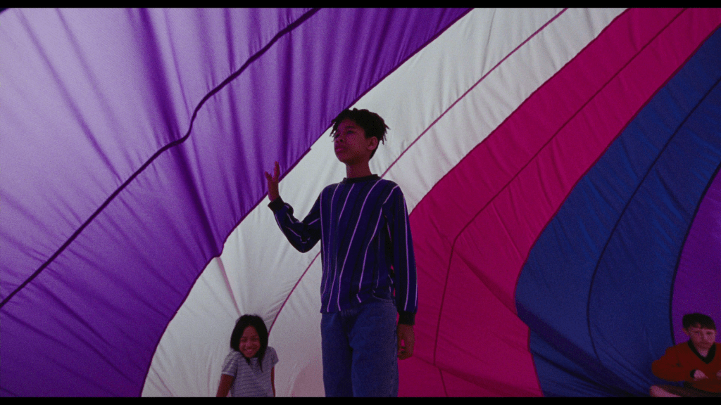 Continuation of the previous shot: Owen dreamily runs his hand along the parachute as he walks under it with two of his classmates visible at the bottom of the frame