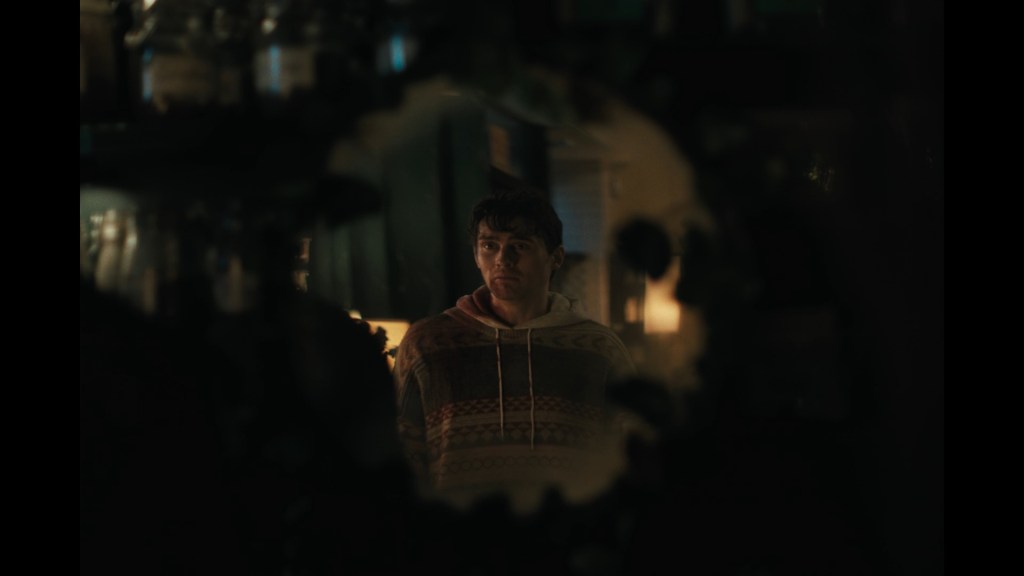 Medium long shot of Bear apologizing to his own reflection in a mirror obscured by shadowy objects such that it looks like an iris shot