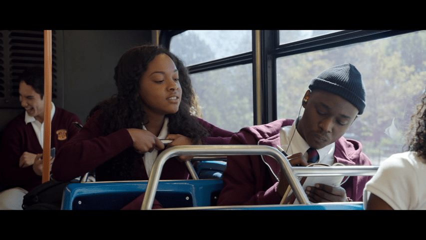 Medium long shot of one of Ashton Sanders's Miles Whittaker drawing in a notebook on a bus while one of his classmates (Penélope de la Rosa) looks over his shoulder from the seat behind him