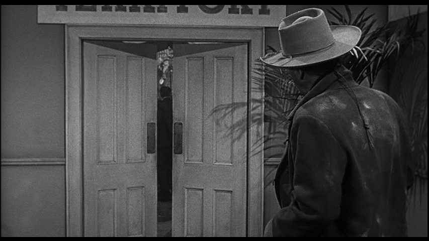 Continuation of the previous shot: Tom Doniphon appears in the right foreground and looks on at the doors with his back to the camera as they swing shut