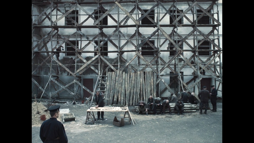 Long shot of a Soviet prison yard depicting prisoners and guards standing or sitting in front of an exterior wall covered by scaffolding that effectively places X's in front of all the windows