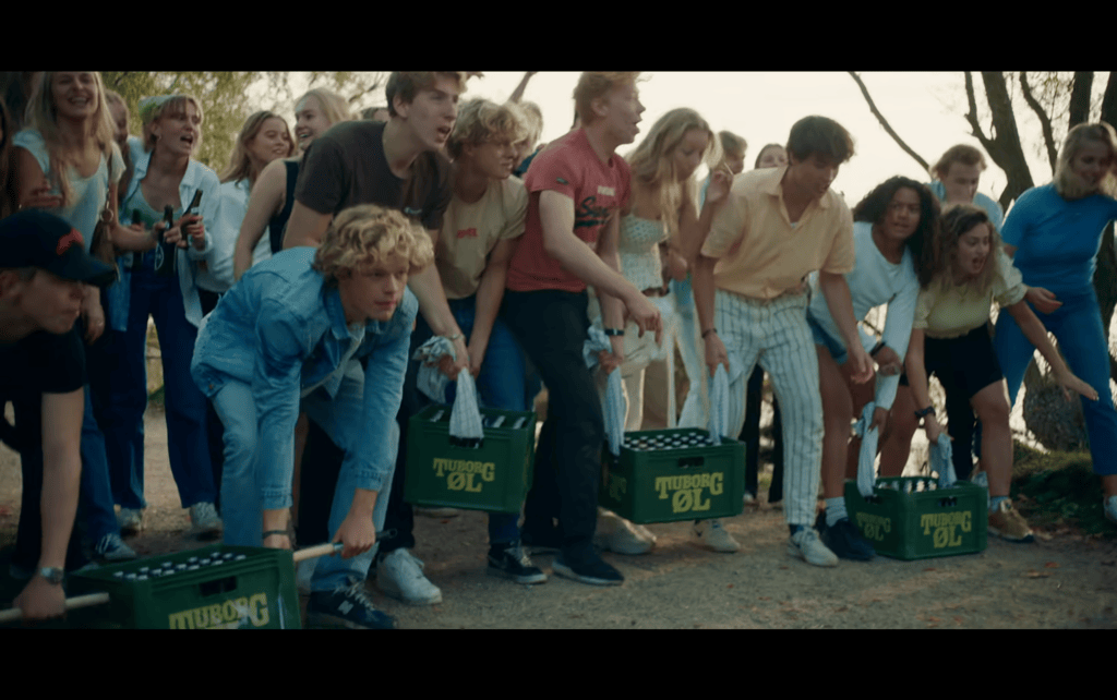 Long shot of a large group of teens assembled at the starting line of a race with cases of beer
