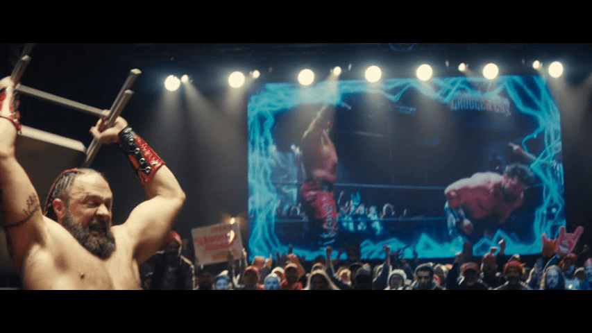 Medium long shot of the wrestler from the previous two images prepares to smash his opponent over the head with a folding chair from the left side of the frame in front of a big screen display showing the same image as a long shot which also includes said opponent
