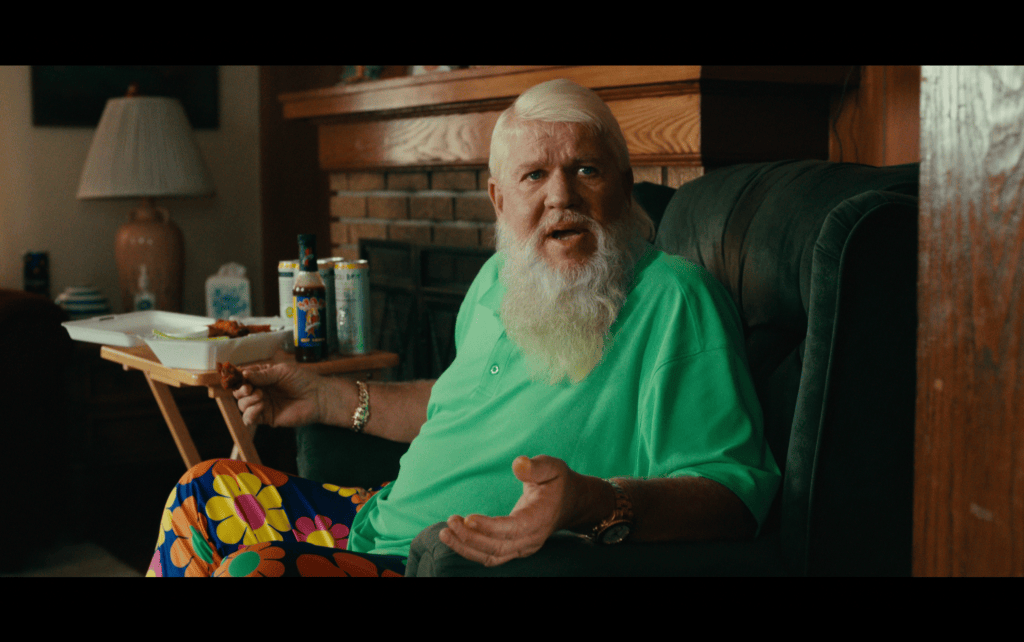 Medium shot of a bearded John Daly in a green golf shirt and garish pants gesturing from a green armchair