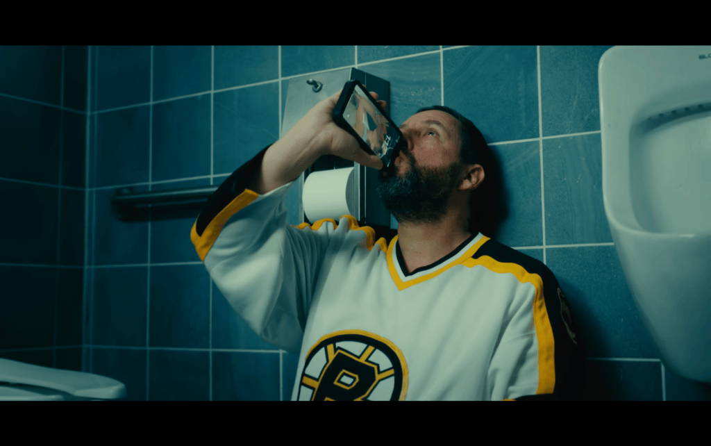 Medium shot of Happy Gilmore on the floor of a bathroom with his trademark Bruins jersey, drinking out of a cellphone