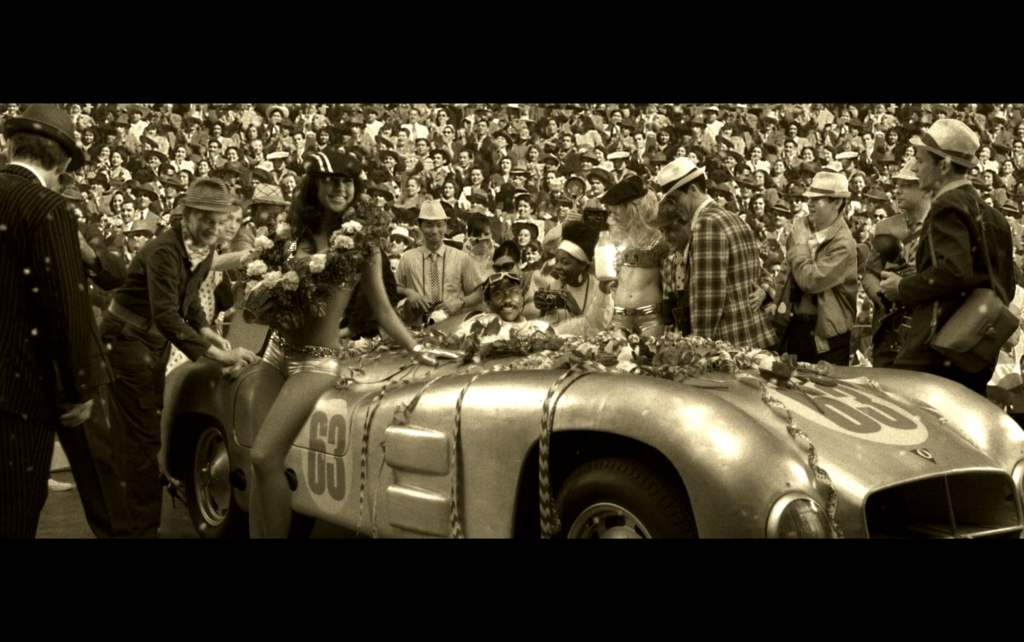 Sepia-toned longshot of the race's winner Ben Burns sitting in his car in front of a crowd celebrating with the traditional bottle of milk
