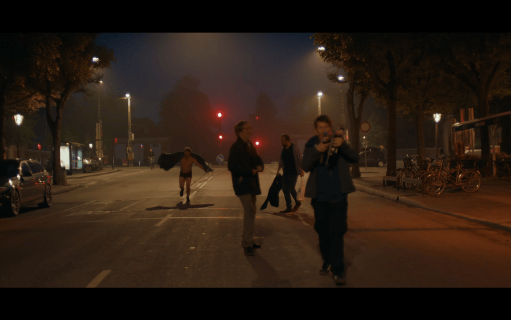 Long shot of four obviously drunken men walking and running through nighttime Copenhagen in the middle of the street