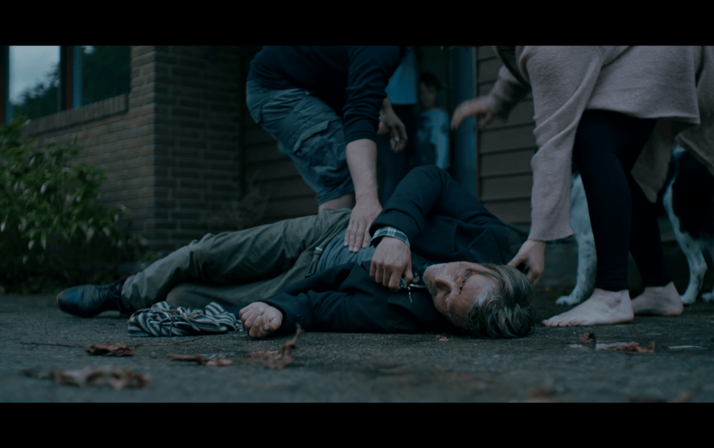 Medium long shot of Martin, who has a nasty-looking cut on his head, passed out in the middle of a driveway as his neighbors attempt to rouse him