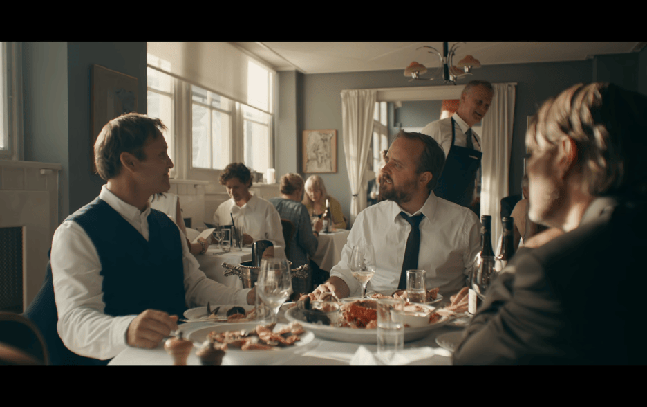 Medium shot of Nikolaj updating Peter and Martin on the state of his marriage as they sit around a table at the restaurant from earlier in the movie