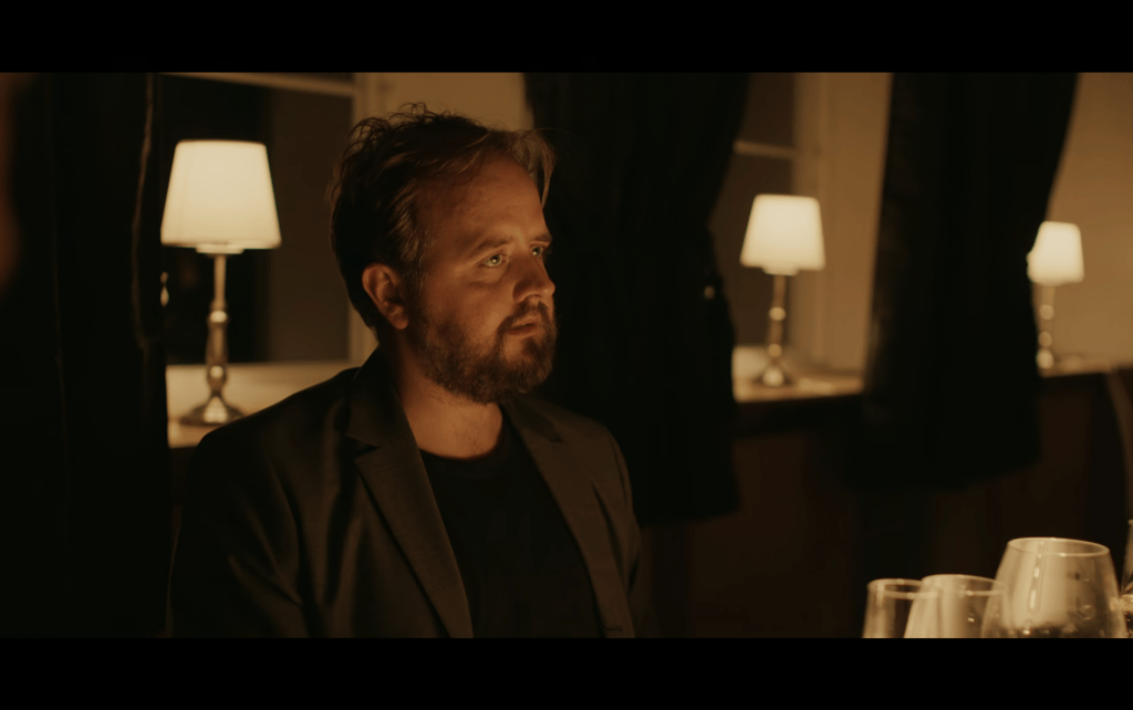 Medium shot of Nikolaj in a dimly-lit restaurant