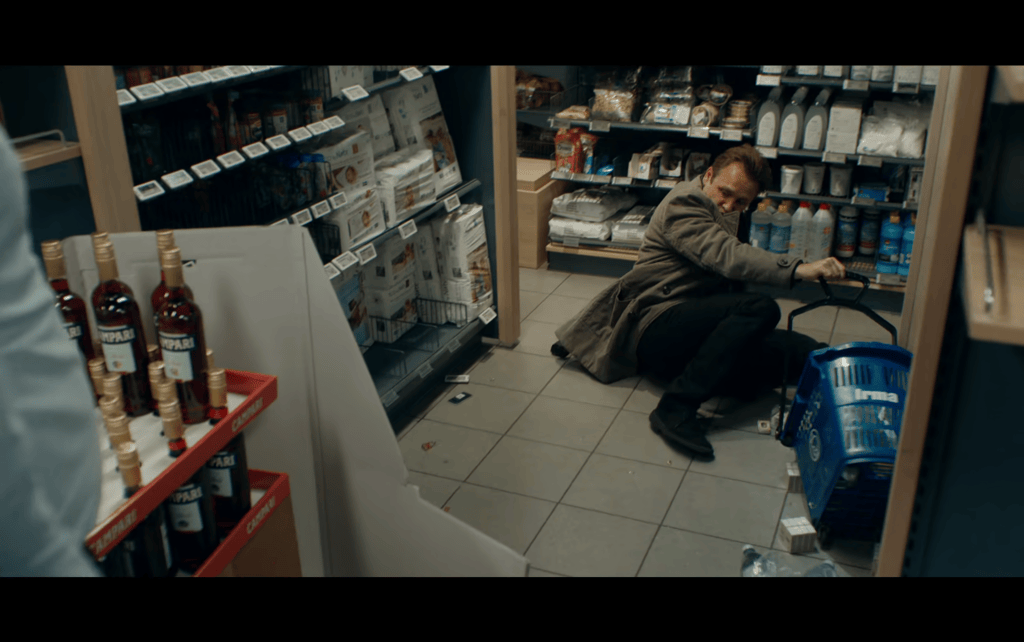 Medium shot of Peter drunkenly sprawled out in the aisle of a grocery store's liquor section where he has fallen, making a mess