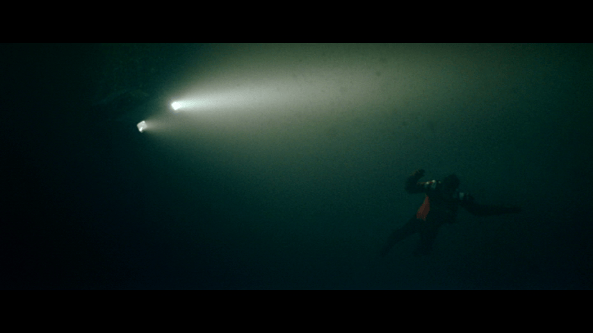 Long shot of Robbie Williams spread eagle in the water lit by the car's headlights